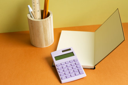 Notebook, calculator, pencil holder on orange desk. yellow wall background. workspace
