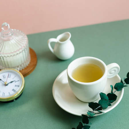 Cup of tea, clock, candle, eucalyptus leaf on green table. pink wall backgroundの写真素材