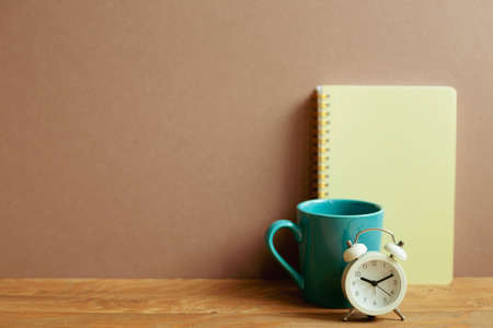 Notebook and mug cup and alarm clock on wooden desk. brown wall background. workspaceの写真素材