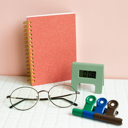 Office desk table with notebook, eyeglasses, clock, clip, supplies on white desk. pink wall...