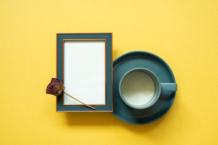 Empty navy photo frame and dry rose flower, cup of milk on yellow background. top view, copy spaceの写真素材