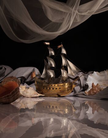 Symbolic composition with a model of an ancient ship, sea shells, waves and clouds on a white glossy tableの写真素材