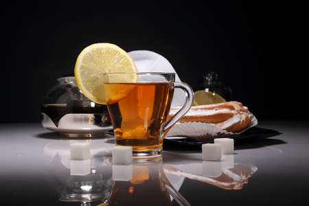 Transparent Cup of tea, lemon, sugar and Eclair on a glossy table and dark backgroundの写真素材
