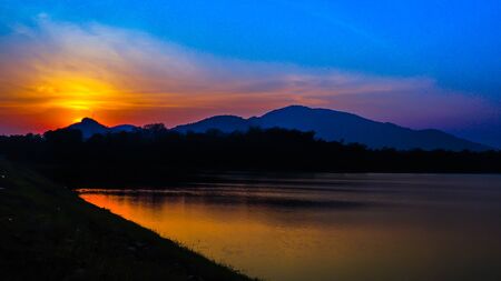 Sunset landscape with orange sky, silhouettes of mountains, hills and trees and lakeの写真素材