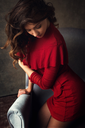 fashion photo of beautiful woman with brunette hair in red dress and black stockings posing  on silver chair on the dark background in studio. Tender atmosphere shoot.の写真素材