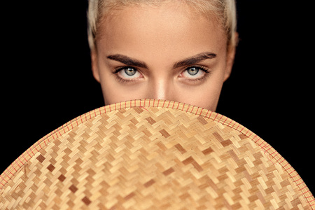 fashion photo of beautiful woman with blonde hair and expressive eyes posing with asian style wicker hat on the black background in studioの写真素材