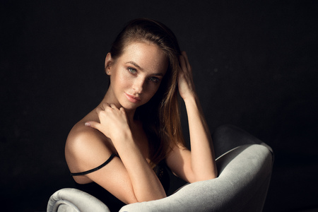 fashion photo of beautiful woman with brunette hair in black dress posing  in sunshine on silver chair on the dark background in studio. Tender atmosphere shoot.の写真素材