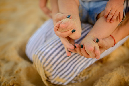 Mother keep in hands the tiny feet of her baby. They seating on the sand of tropicfl beach in Thailandの写真素材