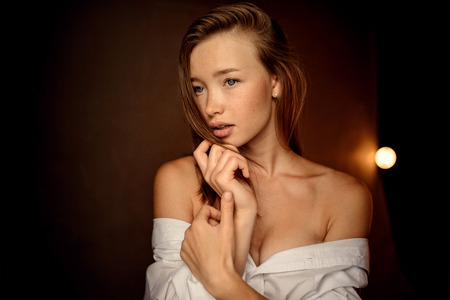 fashion photo of beautiful woman with brunette hair and blue eyes in white shirt posing on dark background in studioの写真素材