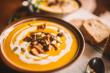 Very appetizing beautiful food photo of tasty autumn coconut pumpkin cream soup bowl with tofu, caramelized onion and seeds. Served with homemade bread on the white towel on wood tableの写真素材