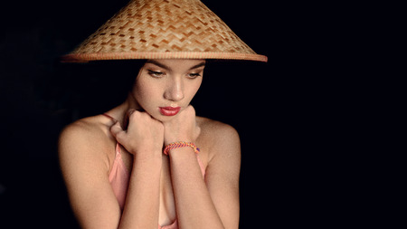 fashion photo of beautiful fit woman with brunette hair in pink bra posing with asian style hat on dark background in studioの写真素材