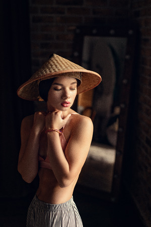 fashion photo of beautiful fit woman with brunette hair in pink bra posing with asian style hat on dark background in studioの写真素材