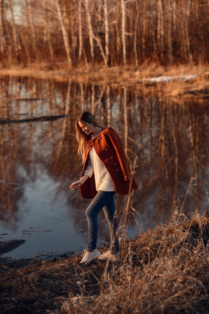 fashion outdoor spring photo of beautiful woman with blonde hair in red coat walking on a sunny day on background of cristal lake and forestの写真素材