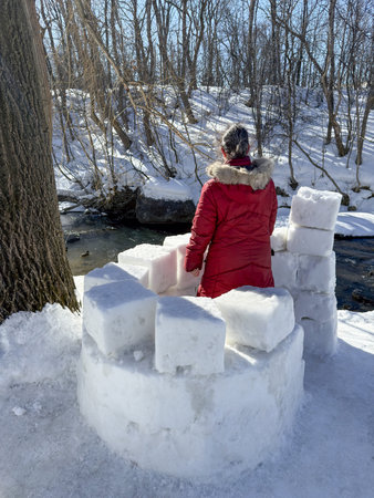 A girl in a red jacket sits on a snow fort and looks at the river. Building a snow structure on the...