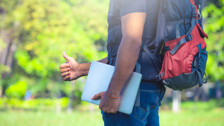 Man with a backpack standing in a park during day . Backpacker holding a laptop. Digital nomad...