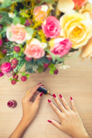 close up of a woman applying red polish on fingernails on table with colorful flowers . Relax , care and beauty conceptの写真素材