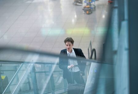 Young businessman on an escalator in the airport, one hand at his rolling case, the other hand in his pocketの写真素材