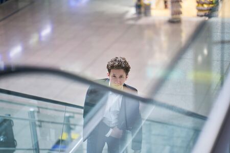 Young handsome smiling businessman inside the airport on an escalator with his rolling suitcaseの写真素材