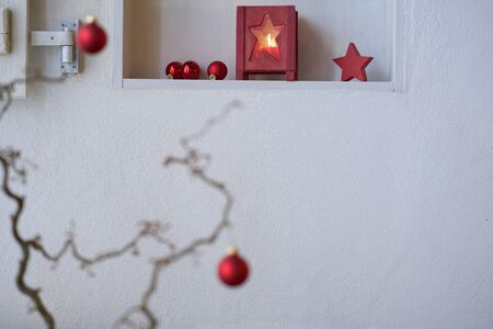 Unsharp red christmas balls at a branch in front of a shelf with red christmas decoration, lantern with candle light, red starの写真素材