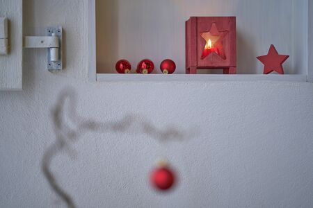 Unsharp red christmas ball at a branch in front of a shelf with red christmas decoration, star, lantern with candle light, white wallの写真素材