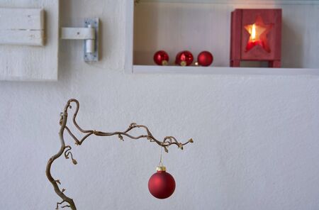 Detail of a red christmas ball at a branch in front of a white wall with candle light and christmas decoration in a shelf with shutterの写真素材
