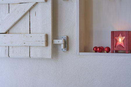 Red christmas decoration in a white shelf in a white wall with a shabby white wooden shutter