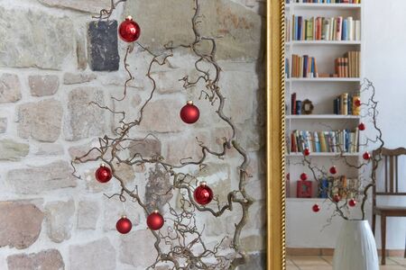 Red christmas decoration at a branch in front of a stone wall, reflections of branch, chair and bookshelf in a mirror with golden frameの写真素材