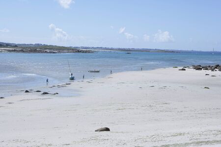 Sunny and relaxing beach with white sand at the coast of Brittany, with small boats and people fishing in the shallow water, pleasant mood, city at the opposite of the bayの写真素材