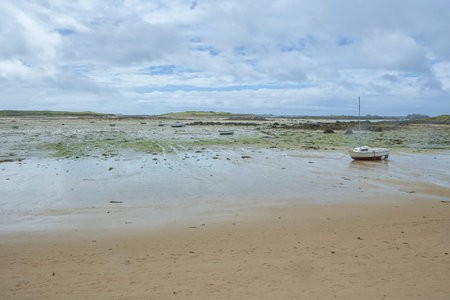 Coast in Brittany at low tide with sand and seaweed and small boats, cloudy blue sky, silent mood