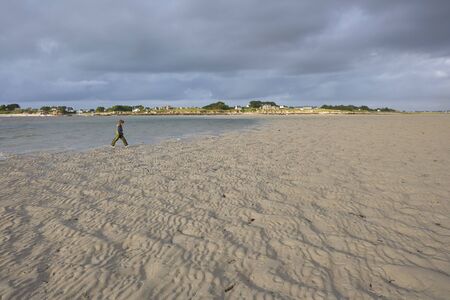 Low tide in a bay in North France, an active boy in rainwear walking on the sand, coastline with houses in the background, cloudy skyの写真素材