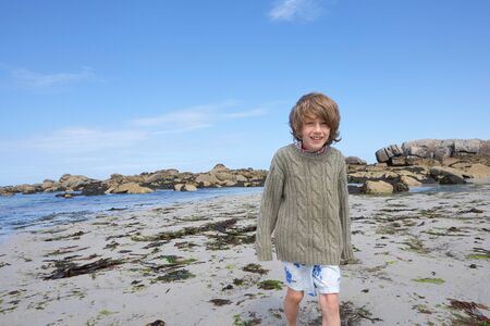Freezing boy at low tide in the sand of a beach with seaweed, water and rocks in the background, summer holiday feelingの写真素材