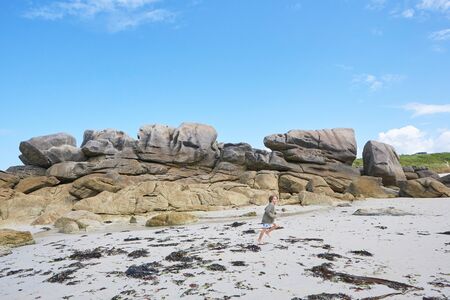 Boy running at ebb in the sand of a bay in Brittany, rock formation in the background, blue summer sky, holiday feelingの写真素材