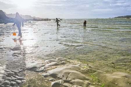 Mother in the foreground and two children in rainwear playing in the shallow water of the sea at a beach, backlightの写真素材