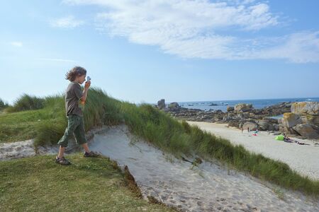 Interested boy concentrating on taking a picture, standing on a dune on a sunny and windy summer day on a beach in Brittanyの写真素材