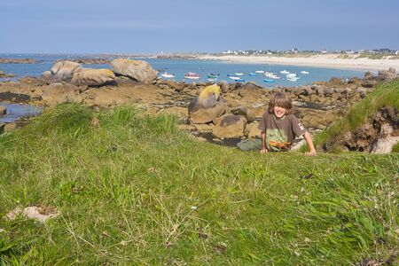 Cheerful boy in the grass at a beach in summer, looking into the camera, in the background small boats in the water and a coastline with housesの写真素材