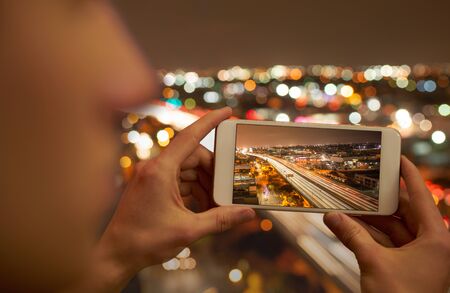 Young man holding a mobile phone taking a picture of an urban situation, unsharp city lights in the backgroundの写真素材