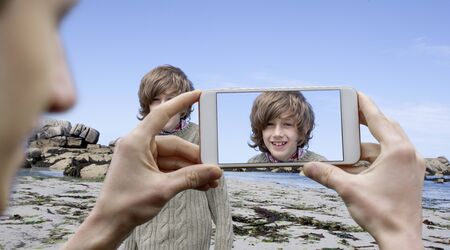 Young man taking a holiday picture with a mobile phone of a boy at a summer beachの写真素材