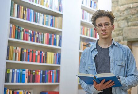 Thoughtful young man with a book in his hand standing in the library of a loft with a stone wall in the background.の写真素材