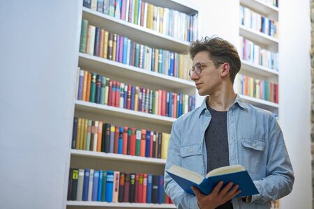 Portrait of a young man looking thoughtful to his right side with a book in his hands, big bookshelves in the background.の写真素材