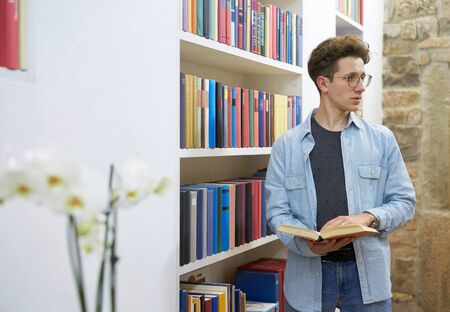 Young student with glasses in library looking to his left side and holding a book in his hands.の写真素材