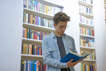 Young handsome man reading a book in a library with big bookshelves in the background.の写真素材