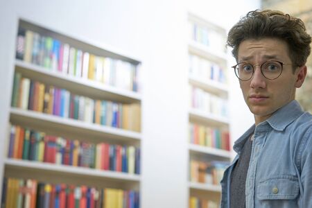 Young student with glasses, looking helpless and overcharged into the camera in library at university.の写真素材