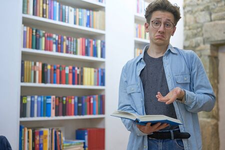 Young studying man with book in his hands standing next to bookshelf and is looking surprised and interrogative in the camera.の写真素材