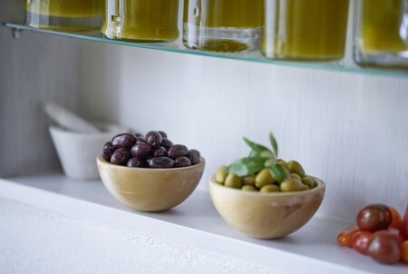 Atmospheric closeup picture of green and black olives in wooden bowls next to blurred white mortar and tomatoes glass bottles with oil content can be seen in the cutの写真素材