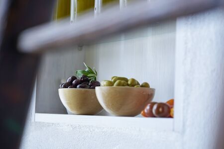 bowls with olives green and brown next to tomatoes stand in the kitchen shelf on the wall in foreground wooden blurred element,photographed in greeceの写真素材
