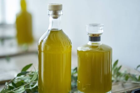 glass bottles filled with a golden liquid oil juice healthy fresh on a rustic wooden piece of furniture olive leaves around them unsharp background. Mediterranean summer feeling wellness relaxed enjoymentの写真素材