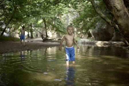 Little boy aged 5 or 6 or 7 standing in shallow water in forest in mediterranean country in summer time sunny relaxed mood teenage boy in backgroundの写真素材