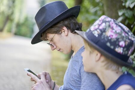 Two relaxed brothers in a park looking at the display of a mobile phone touching another at the shoulderの写真素材