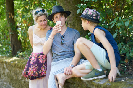 Teenage boy talking into his mobile phone while sitting between brother and mother on a stone wall under trees in a park in summerの写真素材