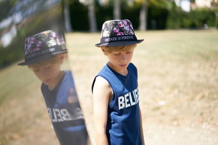 Portrait of a young teenage boy with hat in a summer park reflected on a metal surfaceの写真素材